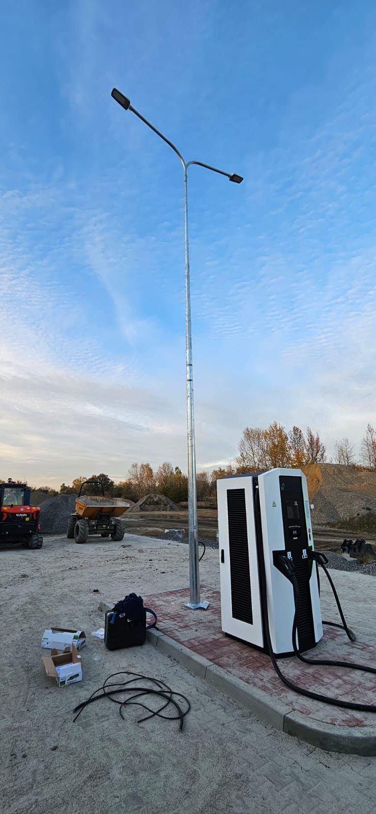 Nowoczesna stacja ładowania pojazdów elektrycznych z dwoma stanowiskami, umieszczona obok metalowego słupa oświetleniowego na placu budowy.