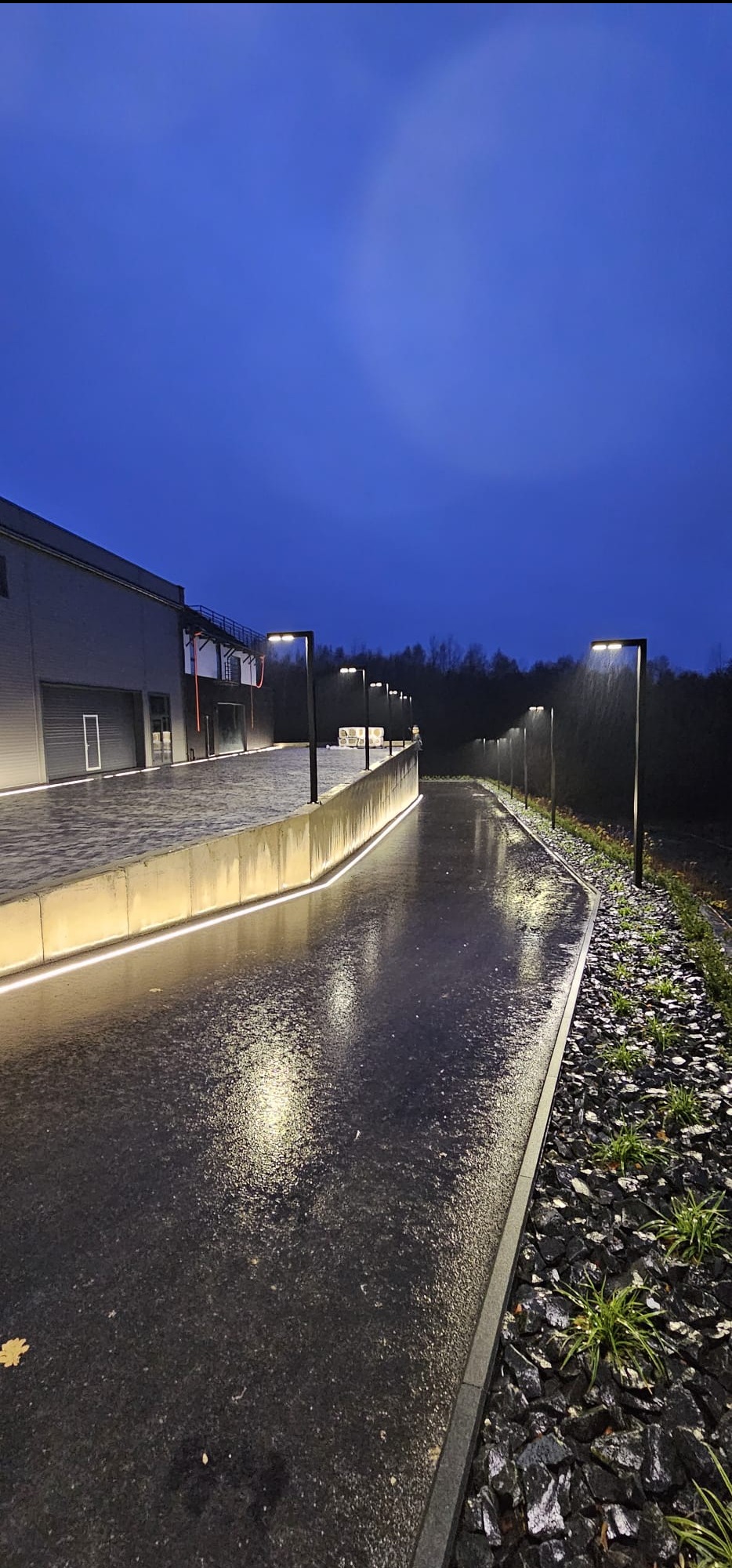 Oświetlona ścieżka wzdłuż budynku z lampami i podświetleniem LED, mokra nawierzchnia odbija światło, kamienny brzeg z roślinami, pochmurne niebo.