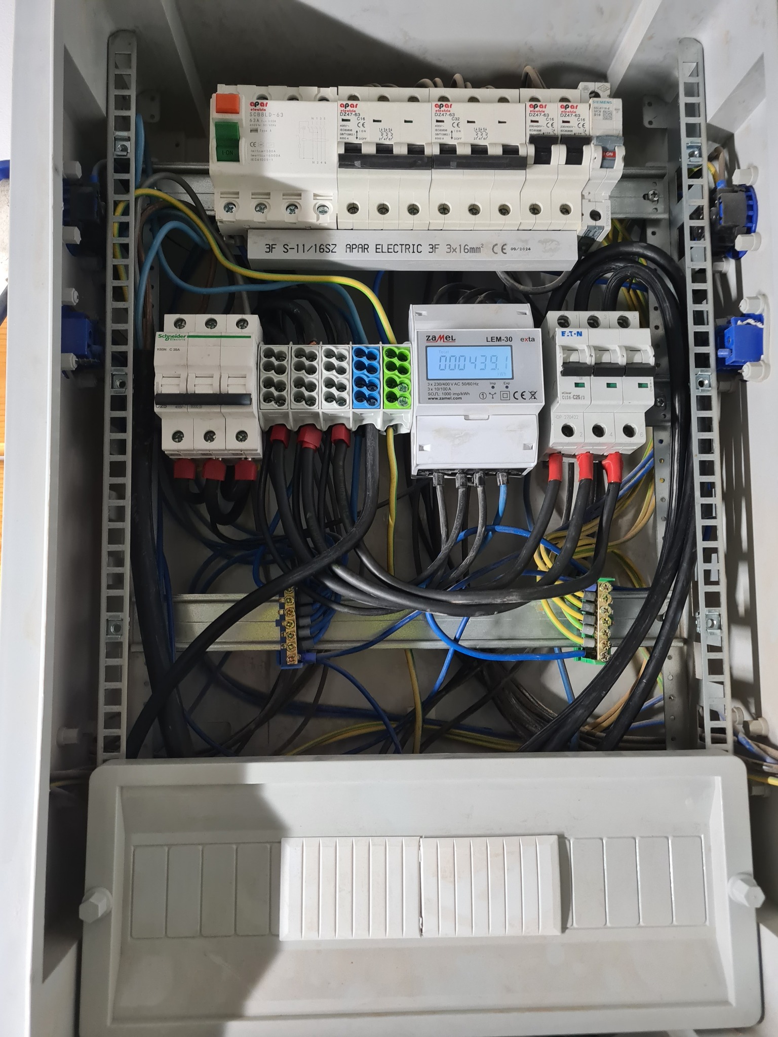 Rozdzielnia elektryczna w obudowie z widocznymi bezpiecznikami, licznikiem energii i przewodami. Układ zasilania w białej skrzynce, widok z góry, detale instalacji.