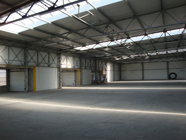 Wnętrze hali magazynowej z betonową posadzką, widoczne bramy segmentowe i konstrukcja stalowa dachu z oknami doświetlającymi, w oddali składowane materiały i opona.