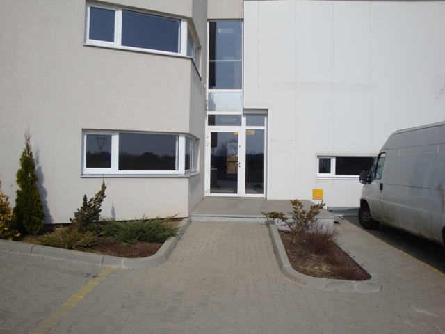 Zewnętrzny widok nowoczesnej hali magazynowej z białymi ścianami, dużymi oknami i przeszklonymi drzwiami wejściowymi, obok zaparkowany biały dostawczy bus, kostka brukowa i małe krzewy przed...