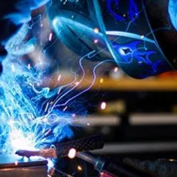Spawanie metalu w warsztacie: zbliżenie na iskrzące się połączenie, spawacz w masce ochronnej z niebieskimi wzorami, rękawice ochronne, widoczne elementy konstrukcji w tle.