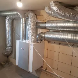 Szare rury wentylacyjne, niektóre owinięte folią aluminiową, połączone z centralą wentylacyjną Wolf w pomieszczeniu z kafelkami i surowym betonem.