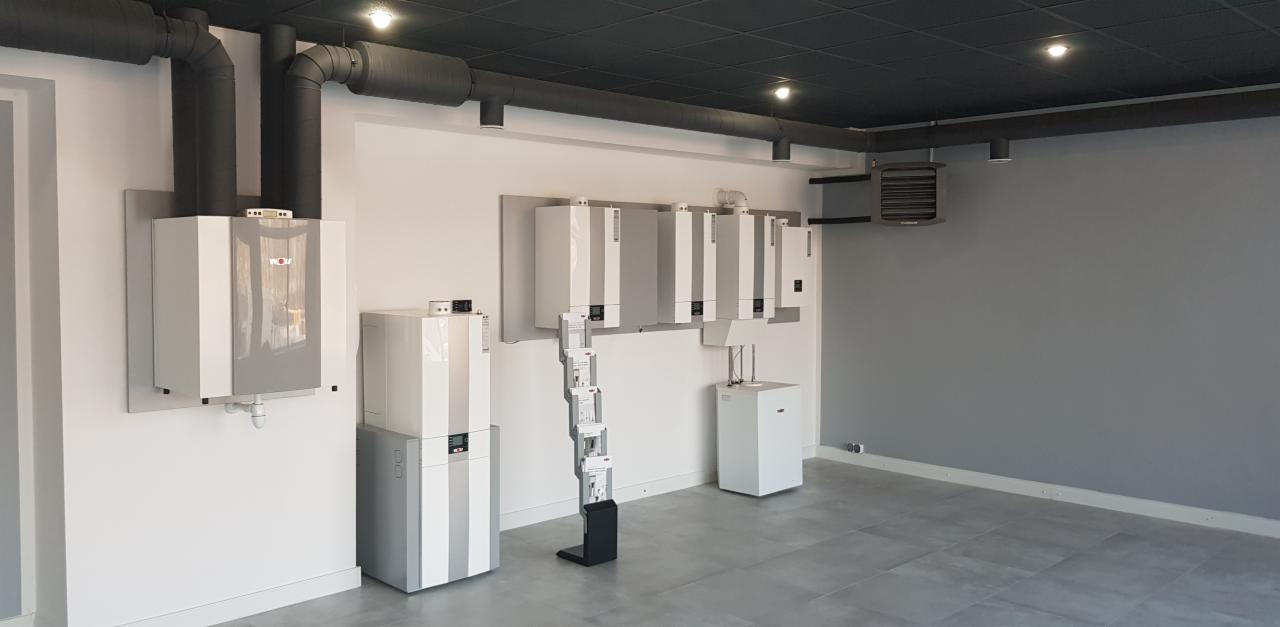 Wnętrze pomieszczenia z zamontowanymi kondensacyjnymi kotłami gazowymi marki Wolf, rurami wentylacyjnymi pod sufitem i stojakiem na ulotki informacyjne.