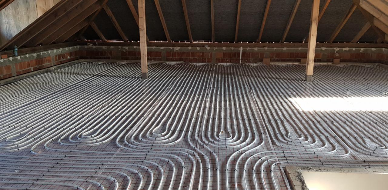 Białe rury ogrzewania podłogowego ułożone w pętle na siatce w surowym stanie poddasza z widoczną konstrukcją dachu i ceglanymi ścianami.