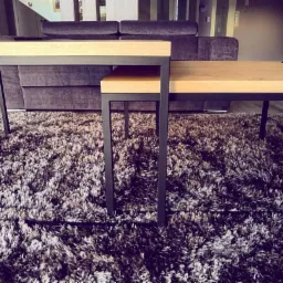 Dwa stoliki kawowe o różnych wysokościach z jasnymi, drewnianymi blatami i ciemnymi, metalowymi nogami, ustawione na puszystym, ciemnym dywanie; w tle widoczna sofa w odcieniach fioletu.