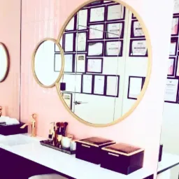 Toaletka z dwoma okrągłymi lustrami w złotych ramach, na blacie czarne pudełka, złote akcesoria kosmetyczne i złota bateria umywalkowa w kształcie ludzkiej figury, w tle różowe płytki i ściana...