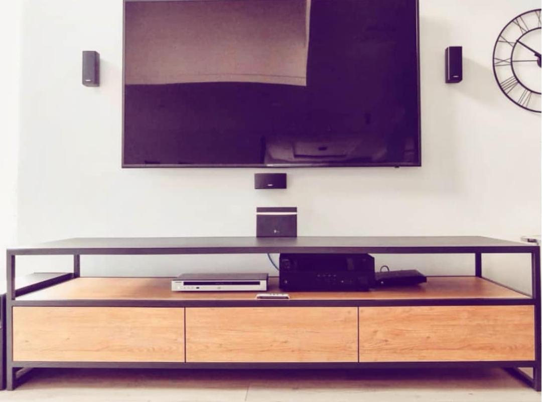 Stylowa szafka RTV na metalowym stelażu z drewnianymi frontami szuflad, pod wiszącym telewizorem i głośnikami, z widocznym zegarem na ścianie.