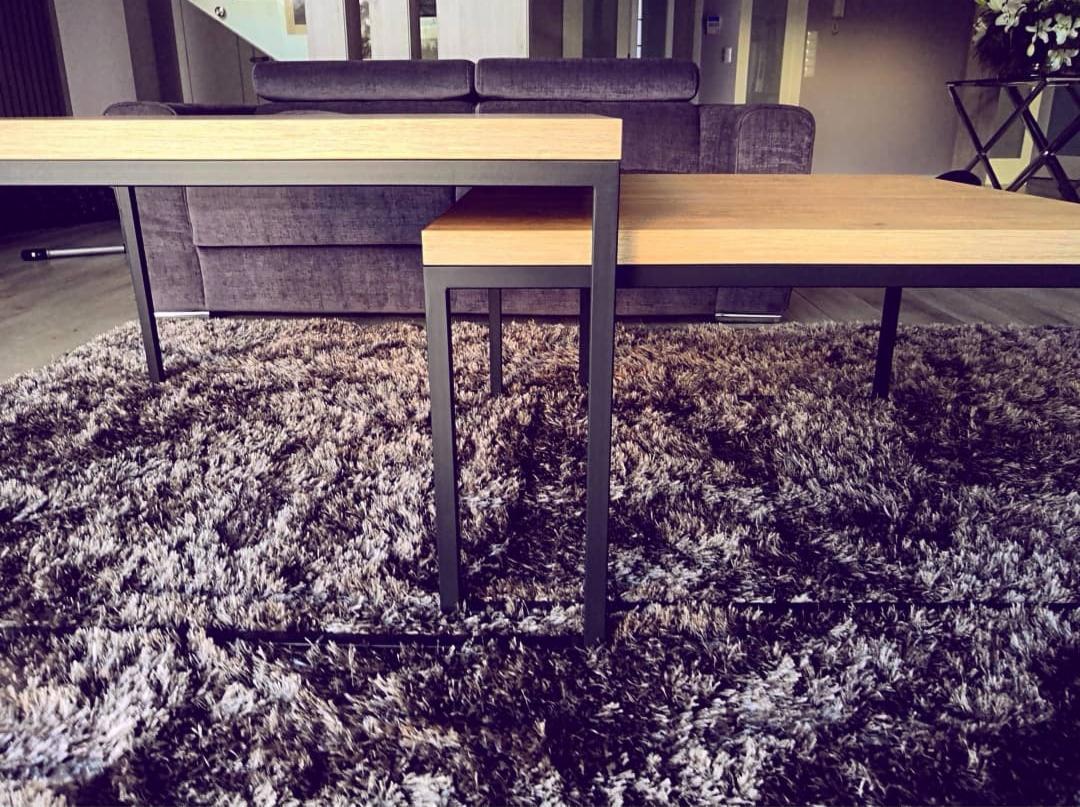 Dwa stoliki kawowe o różnych wysokościach z jasnymi, drewnianymi blatami i ciemnymi, metalowymi nogami, ustawione na puszystym, ciemnym dywanie; w tle widoczna sofa w odcieniach fioletu.