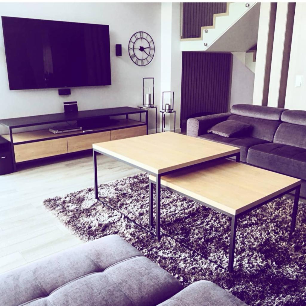 Nowoczesny salon z dwupoziomowym stolikiem kawowym o drewnianym blacie i czarnych metalowych nogach, szarym dywanem shaggy, szarą sofą i szafką RTV pod wiszącym telewizorem.