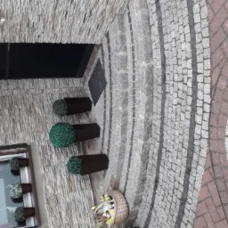 Elewacja budynku wykończona kamieniem z ozdobnymi roślinami w donicach i koszem z kwiatami przy wejściu, obok ułożona kostka brukowa w różnych kolorach tworząca wzór.