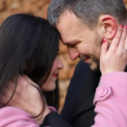 Zakochana para dotyka się czołami na tle ceglanej ściany, kobieta w różowym płaszczu z pierścionkiem na palcu obejmuje twarz mężczyzny w czarnym płaszczu.