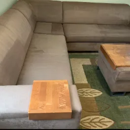 Duży, narożny, brązowy tapicerowany zestaw wypoczynkowy z widocznymi śladami po czyszczeniu, z drewnianymi podłokietnikami i stolikiem na tle dywanu w liście.