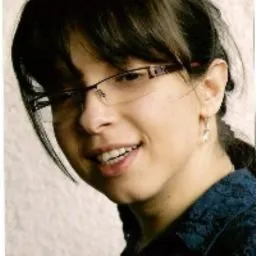 Portret uśmiechniętej kobiety w okularach z ciemnymi włosami splecionymi w warkocz, ubrana w niebieską bluzkę z delikatnym wzorem, na rozmytym, jasnym tle.