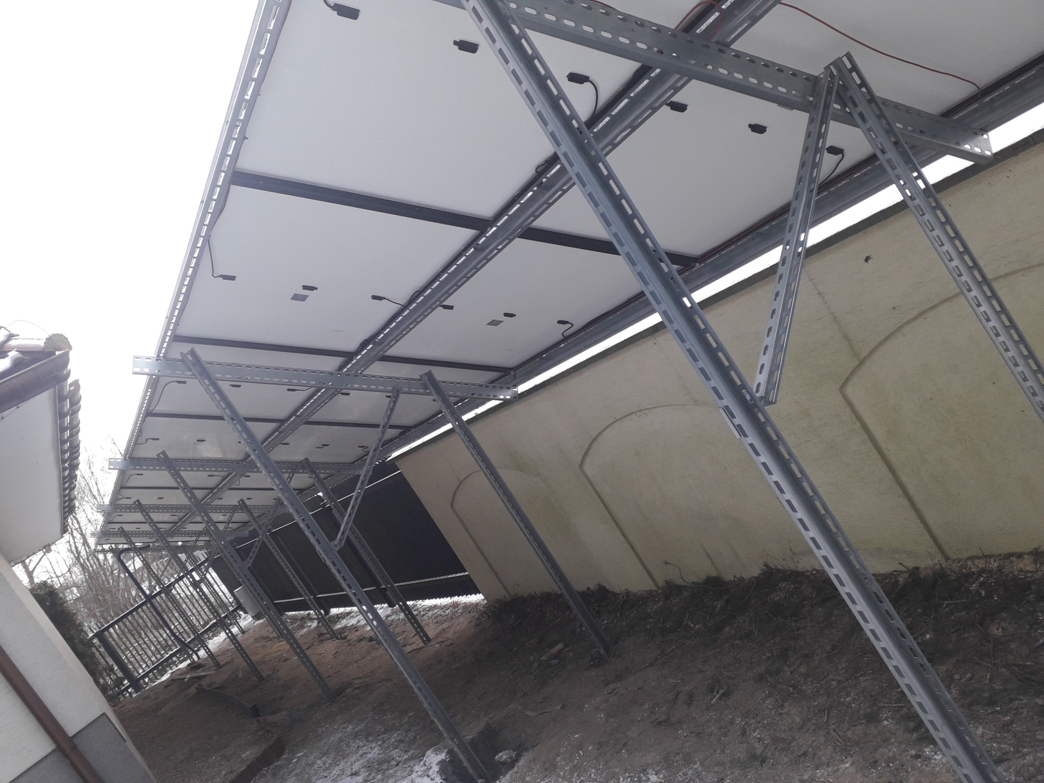 Montaż paneli słonecznych na metalowej konstrukcji, widok od dołu, z ziemi, z fragmentem ściany w tle.