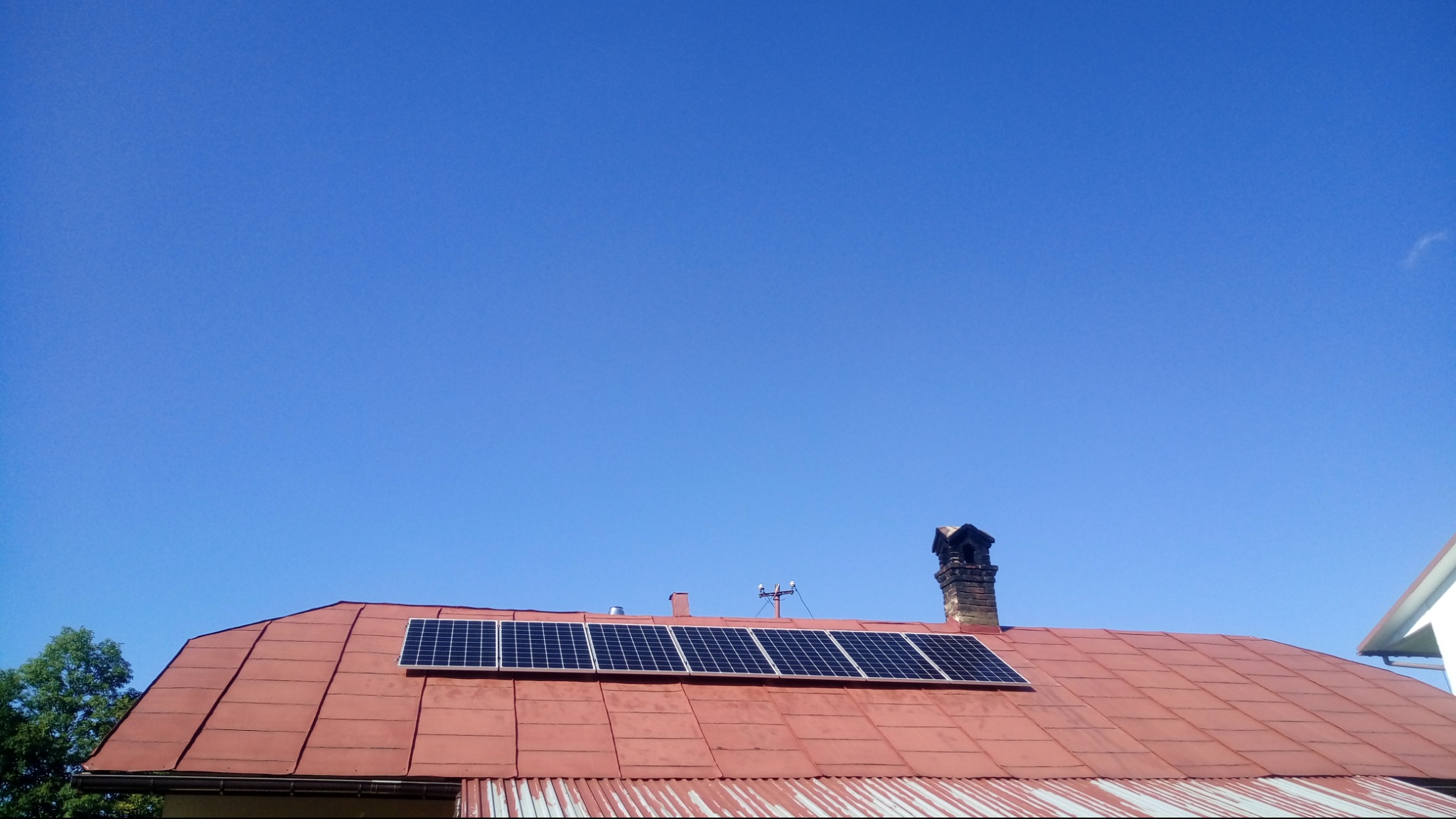 Czerwony dach z blachy trapezowej z zamontowanymi panelami słonecznymi w układzie 2x4, widoczne kominy i antena telewizyjna, błękitne niebo w tle.