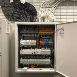 Otwarta szafa sterownicza z widocznymi zabezpieczeniami, przewodami i listwami zaciskowymi, z żółto-czerwonym śrubokrętem leżącym na dole.