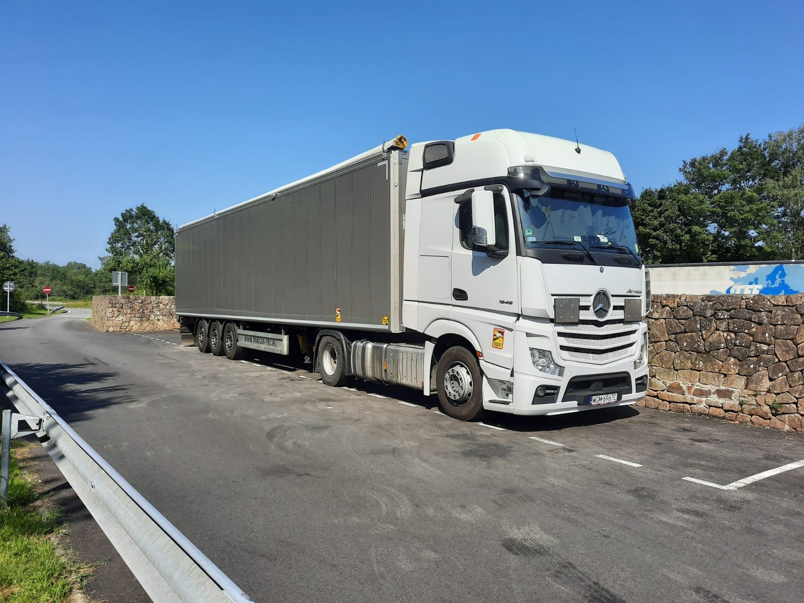 Biała ciężarówka Mercedes Actros z szarą naczepą zaparkowana na parkingu przy kamiennym murku w słoneczny dzień.