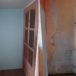 Korytarz w trakcie przygotowania do prac wykończeniowych: ściana pomalowana na błękitno sąsiaduje z fragmentem ściany z resztkami starej, odchodzącej tapety w odcieniach pomarańczu i beżu, widoczne...