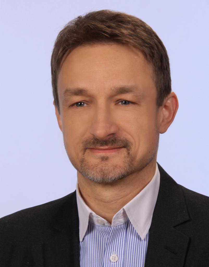 Portret mężczyzny w garniturze i koszuli w paski, z krótką brodą i wąsami, na jasnoniebieskim tle.