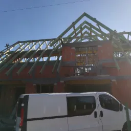 Budowa domu z czerwonej cegły: widoczne ściany w trakcie wznoszenia i drewniana konstrukcja dachu, na pierwszym planie biały samochód dostawczy.