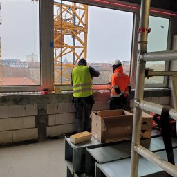 Usługi Ogólnobudowlane Suchecki Dariusz - Dwóch pracowników w odblaskowych kamizelkach montuje okno na tle miejskiego krajobrazu z widocznym żurawiem budowlanym, w pomieszczeniu widoczne elementy instalacji wentylacyjnej i rusztowanie.