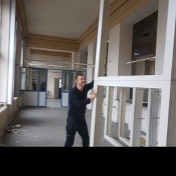 Usługi Ogólnobudowlane Suchecki Dariusz - Uśmiechnięty mężczyzna w czarnym stroju, trzymający białą ramę okienną w surowym wnętrzu z widoczną podłogą w trakcie remontu i innymi oknami w tle.