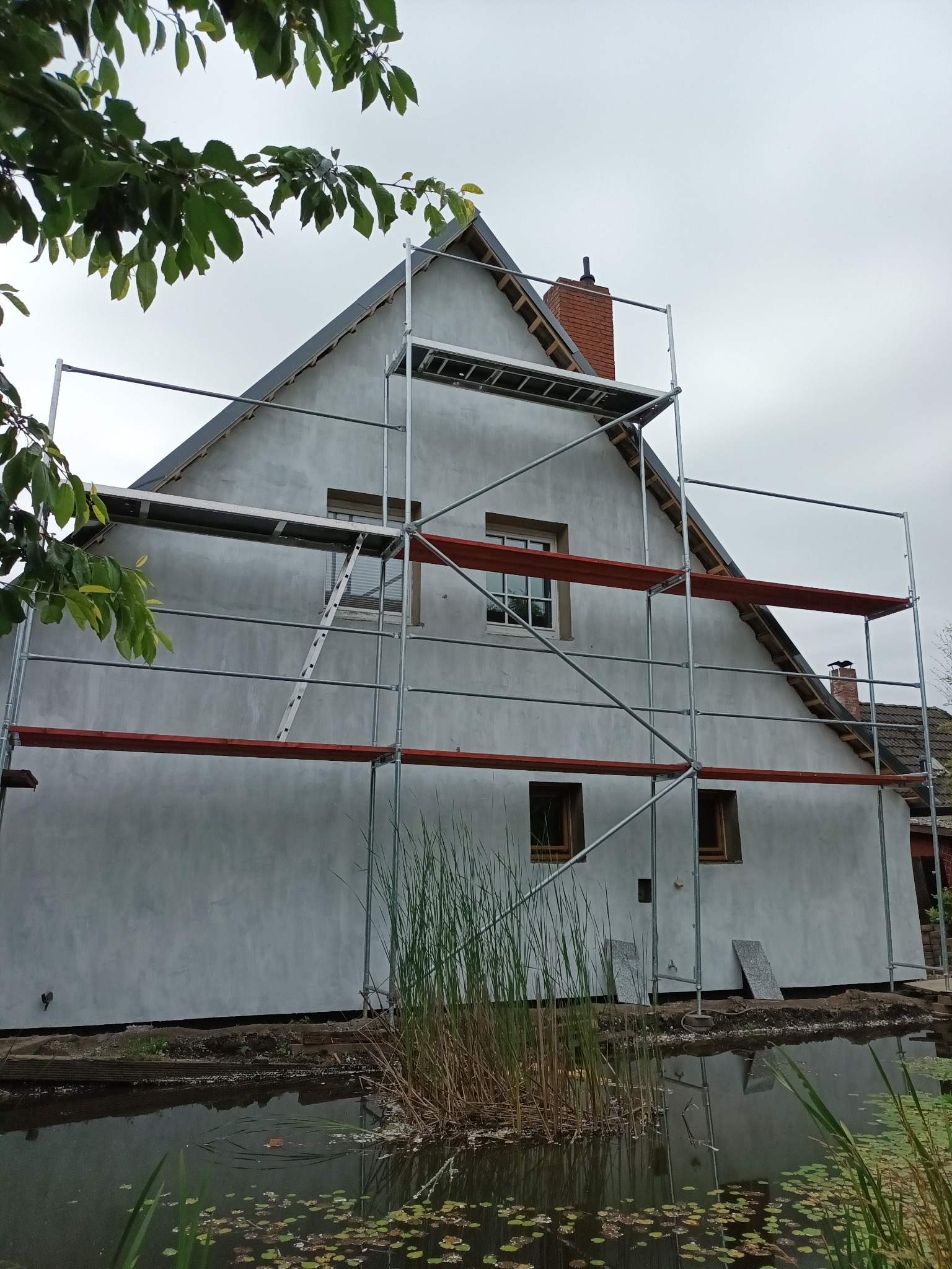 Dom jednorodzinny w trakcie wykonywania elewacji, widoczne rusztowanie, tynk w szarym odcieniu, okna z drewnianą ramą, w tle staw z roślinnością wodną.