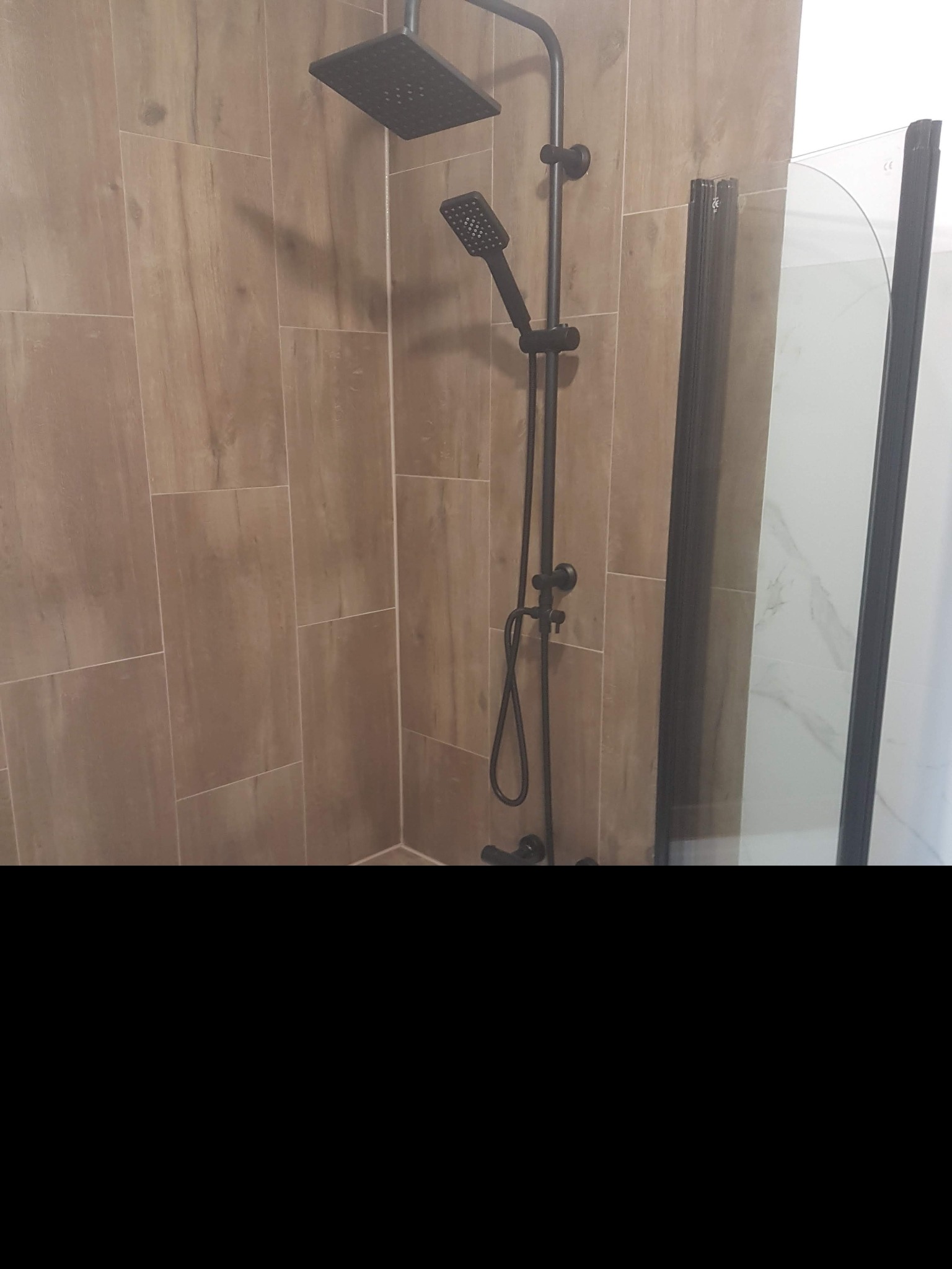 Nowoczesny zestaw prysznicowy w kolorze czarnym, zamontowany na ścianie wyłożonej płytkami imitującymi drewno, obok kabiny prysznicowej z czarną ramą.