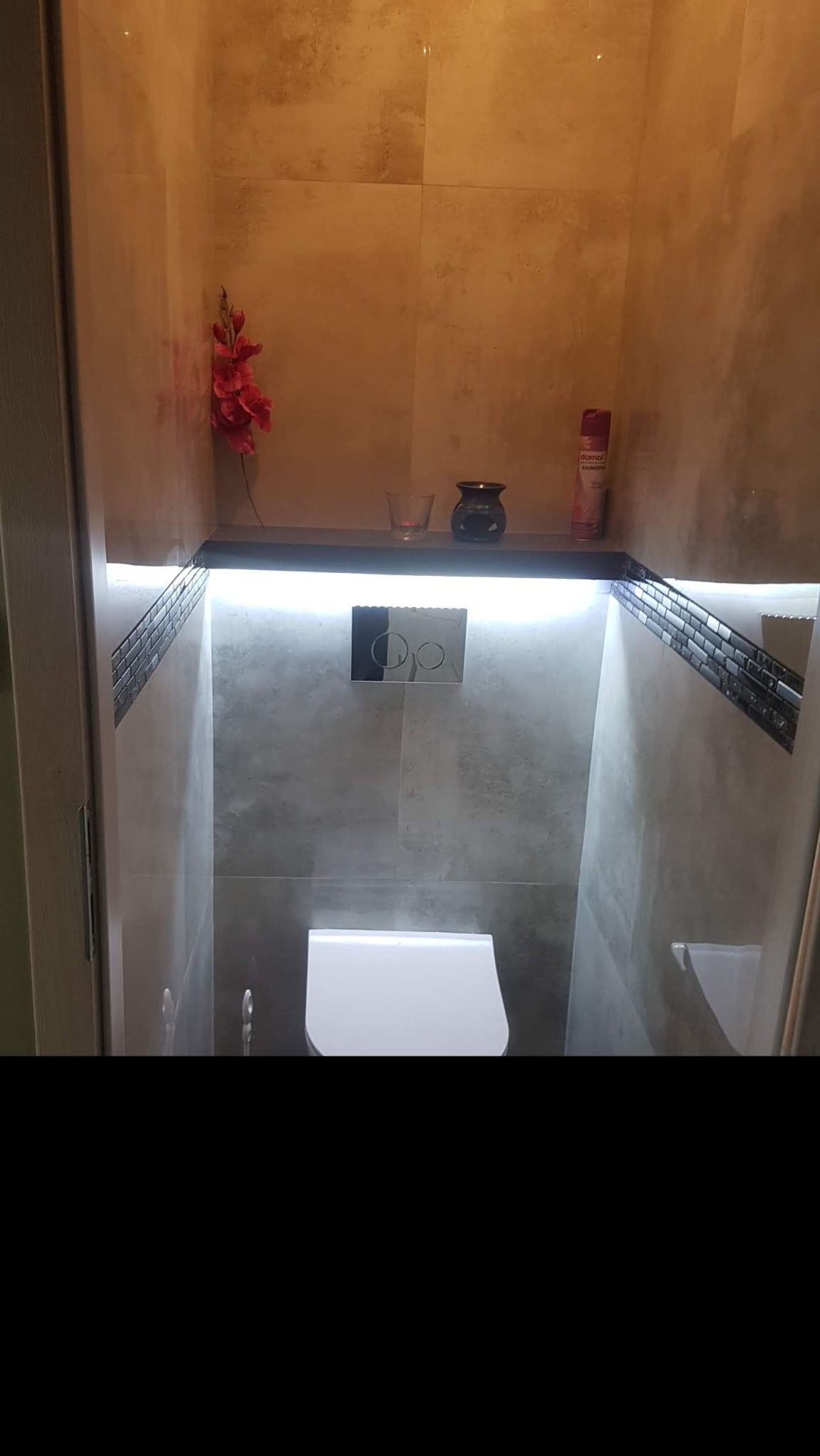 Niewielka, nowoczesna toaleta z płytkami imitującymi kamień, podświetlaną półką z dekoracjami i wiszącą muszlą klozetową.