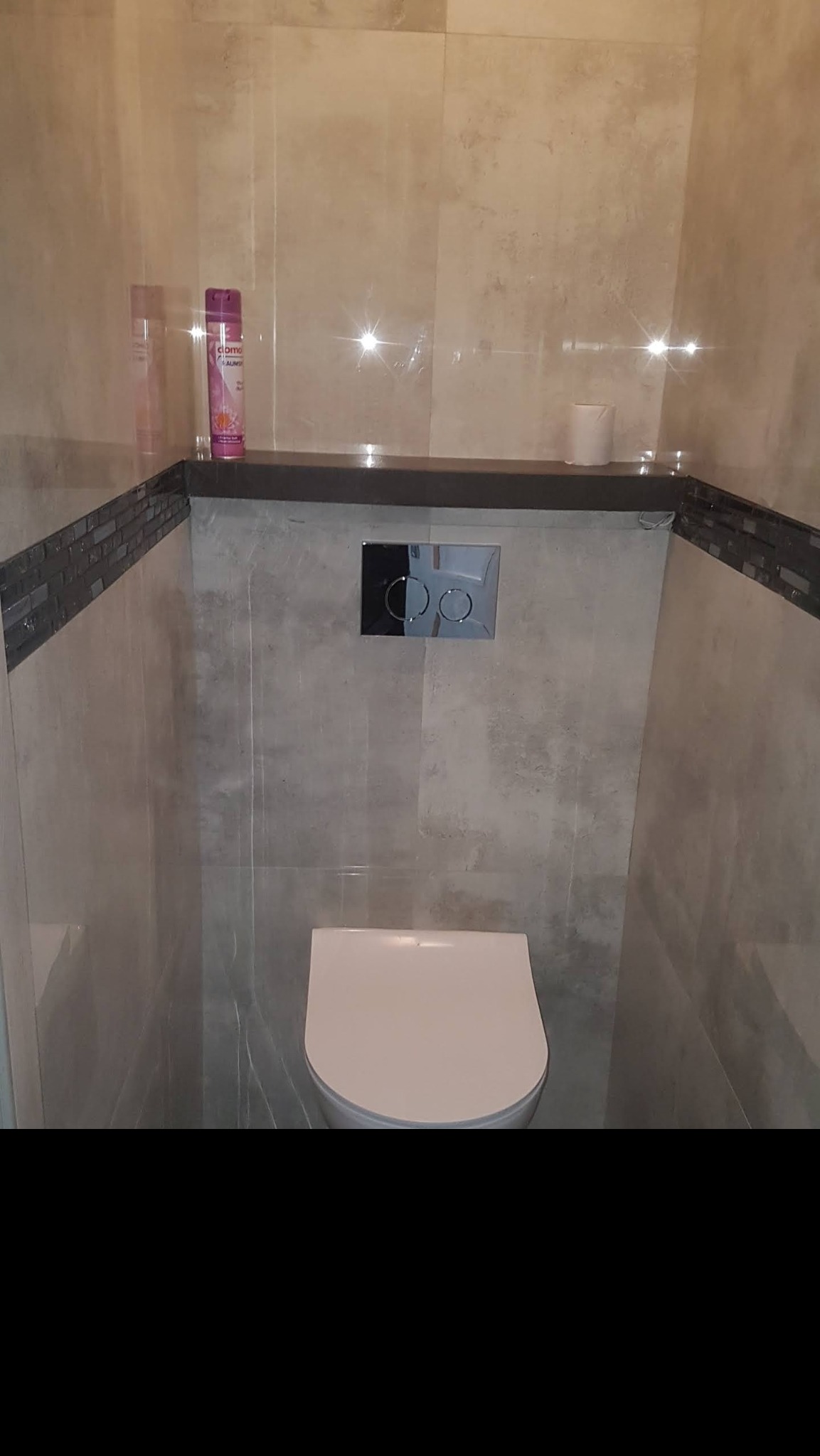 Nowoczesna toaleta w wykończonej łazience z beżowymi płytkami, podwieszanym sedesem, czarną półką i chromowanym przyciskiem spłukującym.