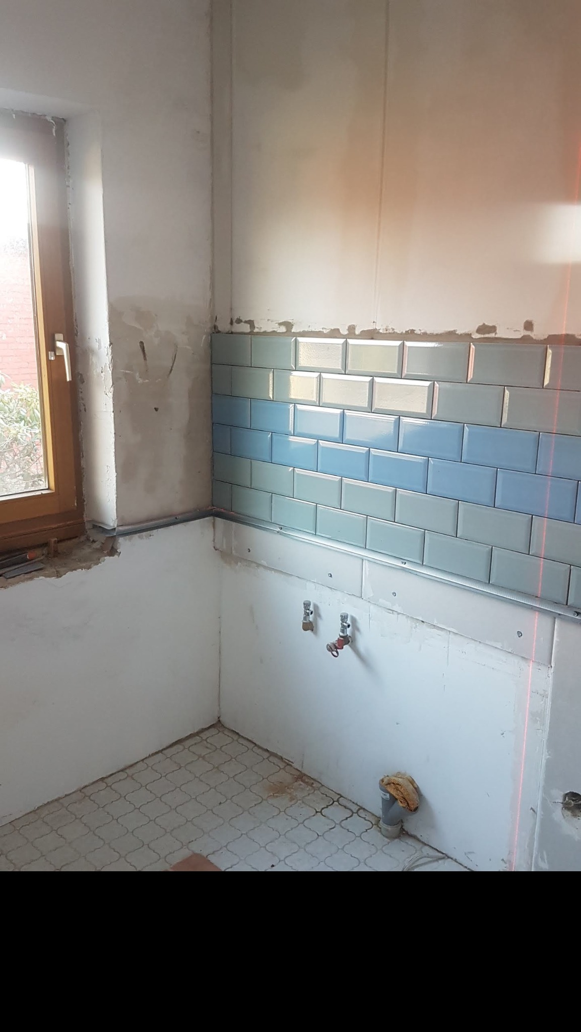 Ściana łazienki w trakcie remontu, częściowo wyłożona błyszczącymi, niebiesko-białymi kafelkami typu metro, widoczne rury, okno oraz laserowy wskaźnik poziomu.