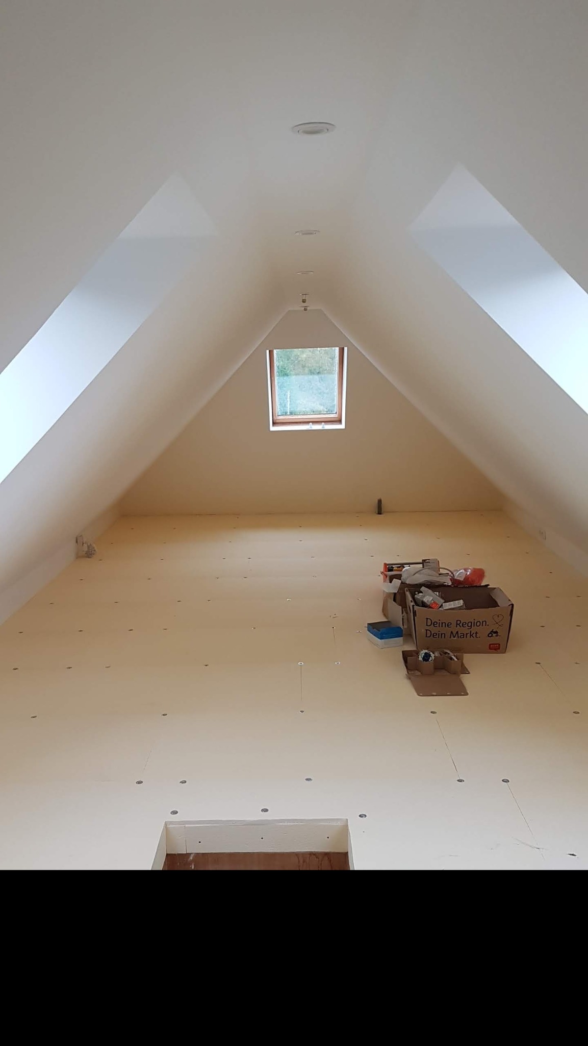 Poddasze w trakcie wykańczania, widoczne płyty podłogowe z otworami, okno dachowe i kartonowe pudła z narzędziami.