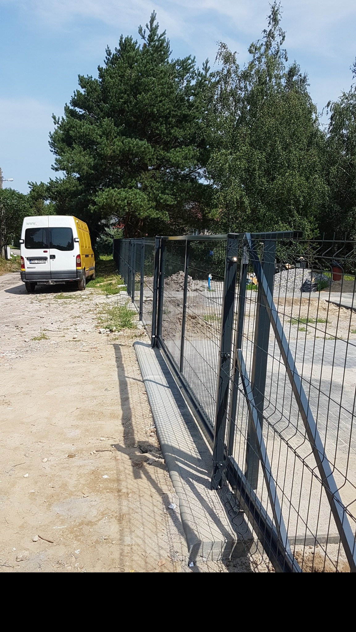 Nowoczesne ogrodzenie panelowe w kolorze grafitowym z przesuwną bramą wjazdową, widok z zewnątrz, obok zaparkowany biało-żółty samochód dostawczy, w tle drzewa.