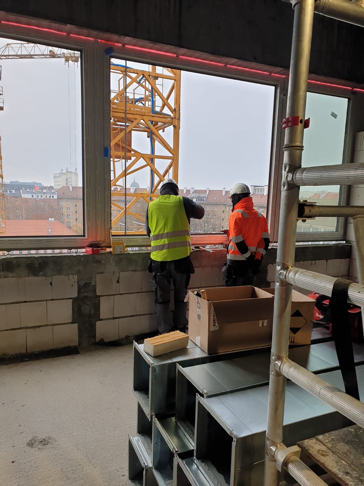 Dwóch pracowników w odblaskowych kamizelkach montuje okno na tle miejskiego krajobrazu z widocznym żurawiem budowlanym, w pomieszczeniu widoczne elementy instalacji wentylacyjnej i rusztowanie.