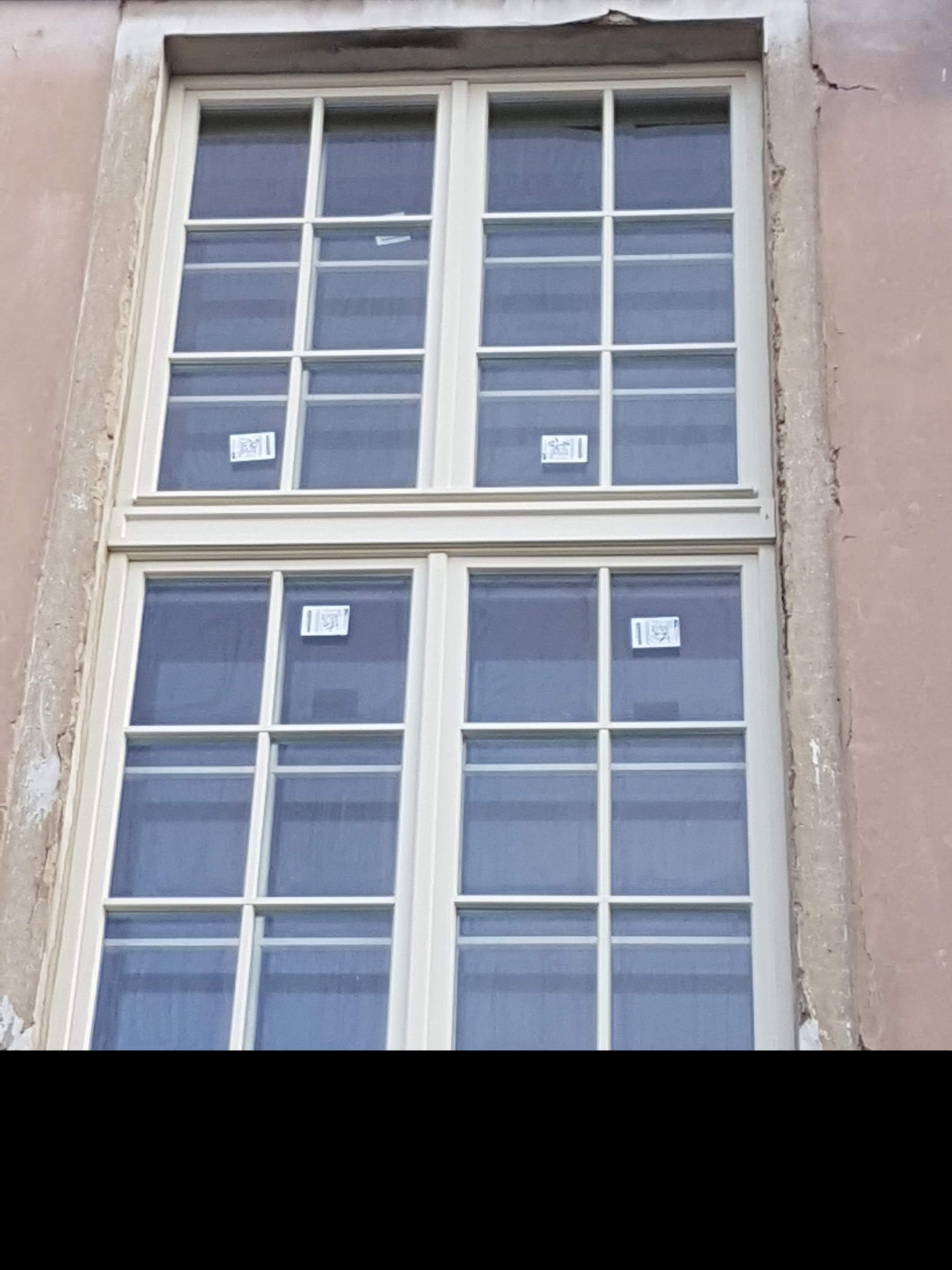 Dwuskrzydłowe, białe okno z kratownicą, z naklejonymi kartkami, osadzone w starej, lekko uszkodzonej elewacji budynku.