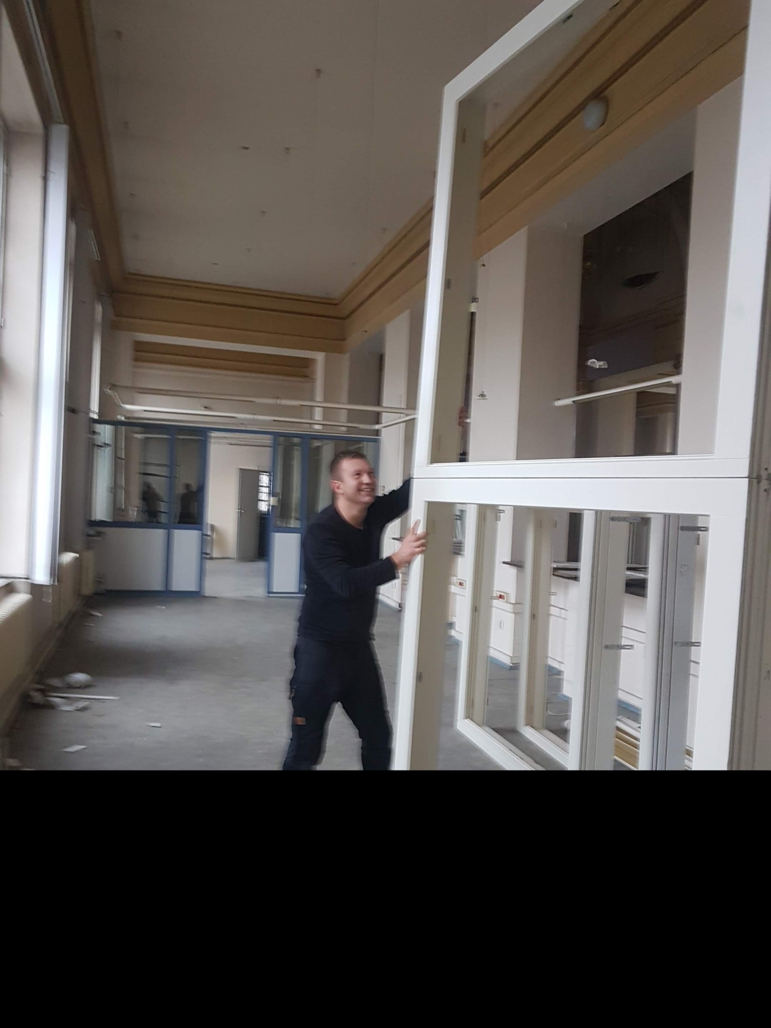 Uśmiechnięty mężczyzna w czarnym stroju, trzymający białą ramę okienną w surowym wnętrzu z widoczną podłogą w trakcie remontu i innymi oknami w tle.