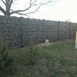 Długi, betonowy płot z dekoracyjnym wzorem imitującym kamień, pomalowany na ciemny kolor z jaśniejszymi akcentami, widoczny z perspektywy trawnika z młodymi iglakami i pniem ściętego drzewa.