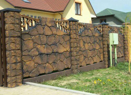Elewacja ogrodzenia z betonu architektonicznego imitującego kamień, z ozdobnymi metalowymi elementami, na tle zielonego trawnika i domów.
