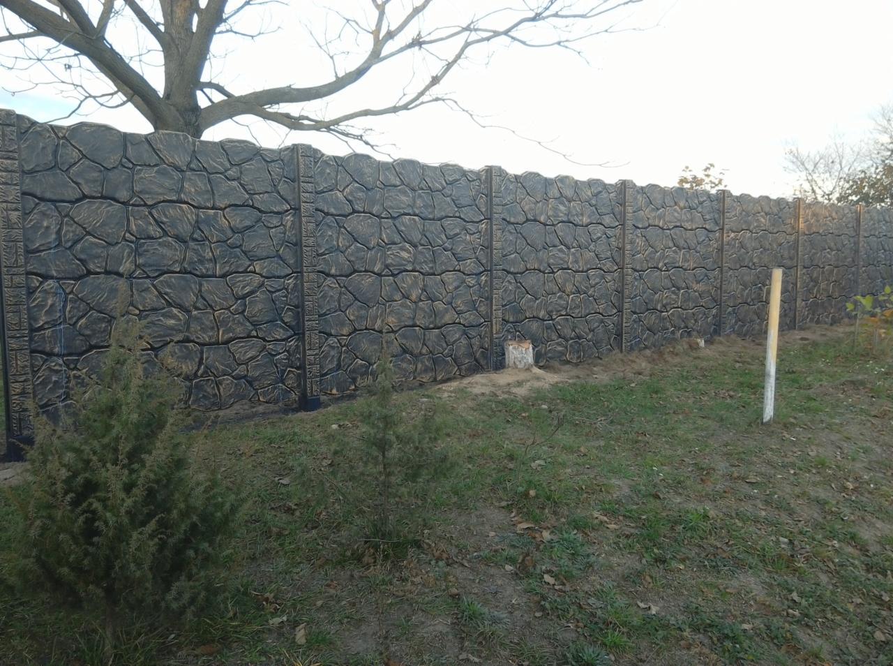 Długi, betonowy płot z dekoracyjnym wzorem imitującym kamień, pomalowany na ciemny kolor z jaśniejszymi akcentami, widoczny z perspektywy trawnika z młodymi iglakami i pniem ściętego drzewa.