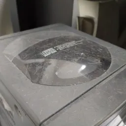 Bardzo zakurzony, szary odtwarzacz CD z widocznymi zarysowaniami i włosami, stojący w zagraconym otoczeniu.