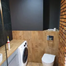 Nowoczesna łazienka z pralką pod blatem, drewnopodobnymi płytkami i ścianą z cegły, oświetlona punktowo.
