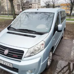 Błękitny van Fiat Scudo na parkingu w pochmurny dzień, widoczne odbicie drzew w kałuży obok pojazdu.