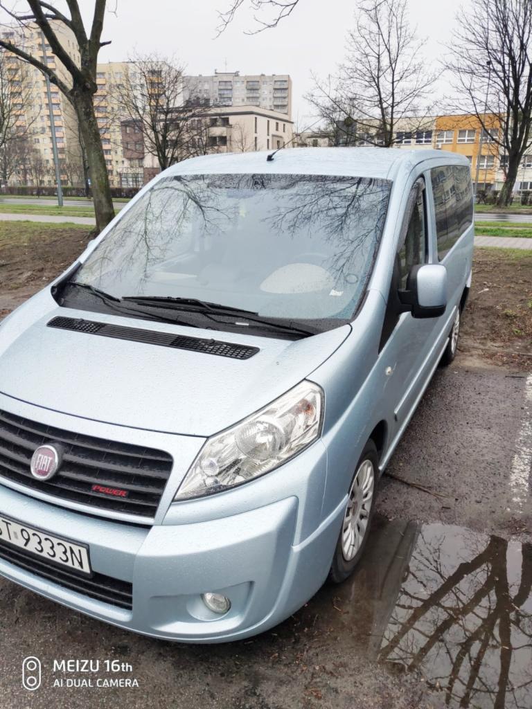 Błękitny van Fiat Scudo na parkingu w pochmurny dzień, widoczne odbicie drzew w kałuży obok pojazdu.