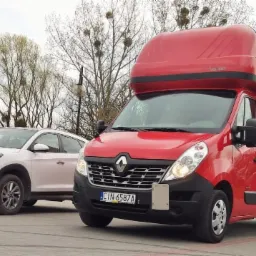 Czerwony bus dostawczy Renault Master z wysoką zabudową i plandeką, zaparkowany obok białego samochodu osobowego Hyundai Tucson na betonowym placu. W tle drzewa bez liści.