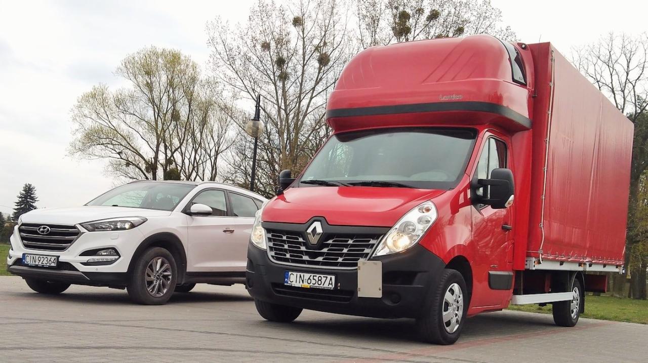 Czerwony bus dostawczy Renault Master z wysoką zabudową i plandeką, zaparkowany obok białego samochodu osobowego Hyundai Tucson na betonowym placu. W tle drzewa bez liści.