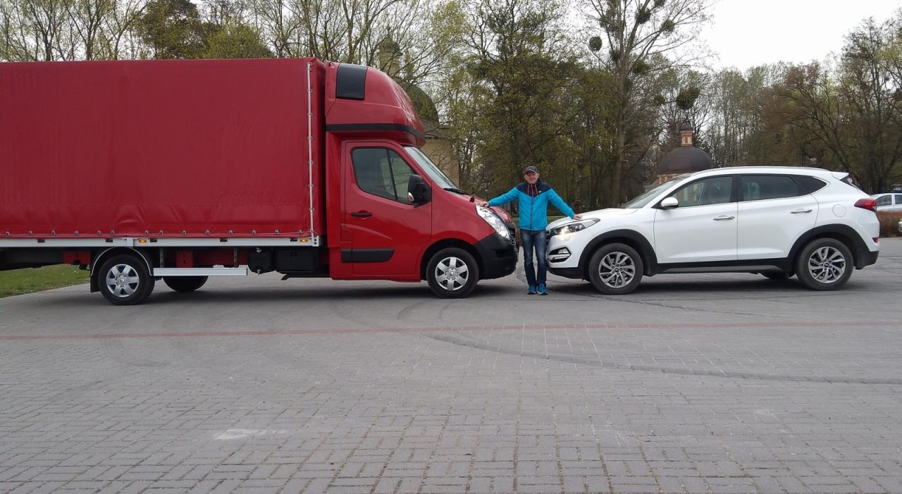 Czerwony bus dostawczy z plandeką i biały samochód osobowy stojące obok siebie na brukowanym placu, osoba opiera się o oba pojazdy.