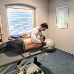 Mężczyzna w białej bluzie wykonuje masaż pleców pacjenta leżącego na profesjonalnym stole do masażu w gabinecie, widoczna mapa kręgosłupa na ścianie i drabinka gimnastyczna.