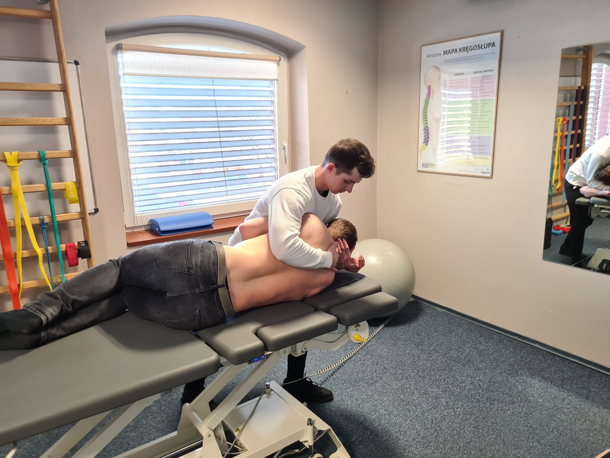 Mężczyzna w białej bluzie wykonuje masaż pleców pacjenta leżącego na profesjonalnym stole do masażu w gabinecie, widoczna mapa kręgosłupa na ścianie i drabinka gimnastyczna.