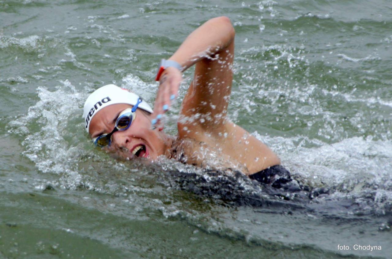 Kobieta w czarnym stroju kąpielowym, czepku i okularach pływackich, płynąca kraulem w wzburzonej wodzie, z widocznymi kroplami wody i ekspresją na twarzy.